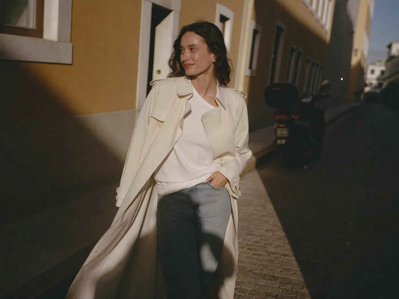 Frau im cremefarbenen Leinen-Trenchcoat in einer sonnigen Altstadtgasse, Hand in der Manteltasche